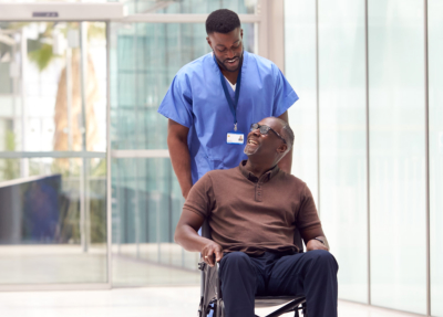 Male Nurse Wearing Scrubs Wheeling Patient In Wheelchair Through Lobby Of Modern Hospital Building